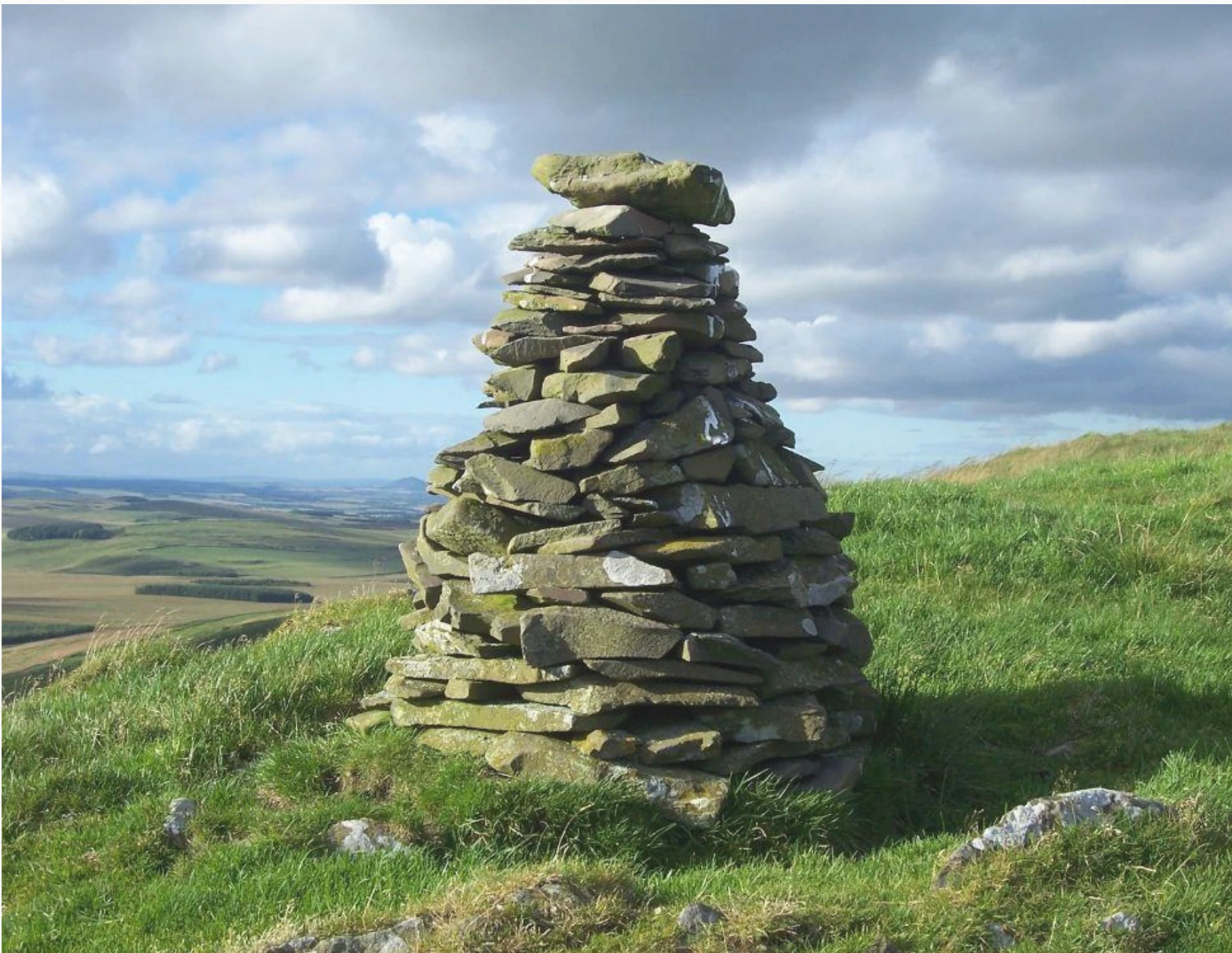 A traditional dry-stone cairn standing on a grassy hillside under a cloudy sky