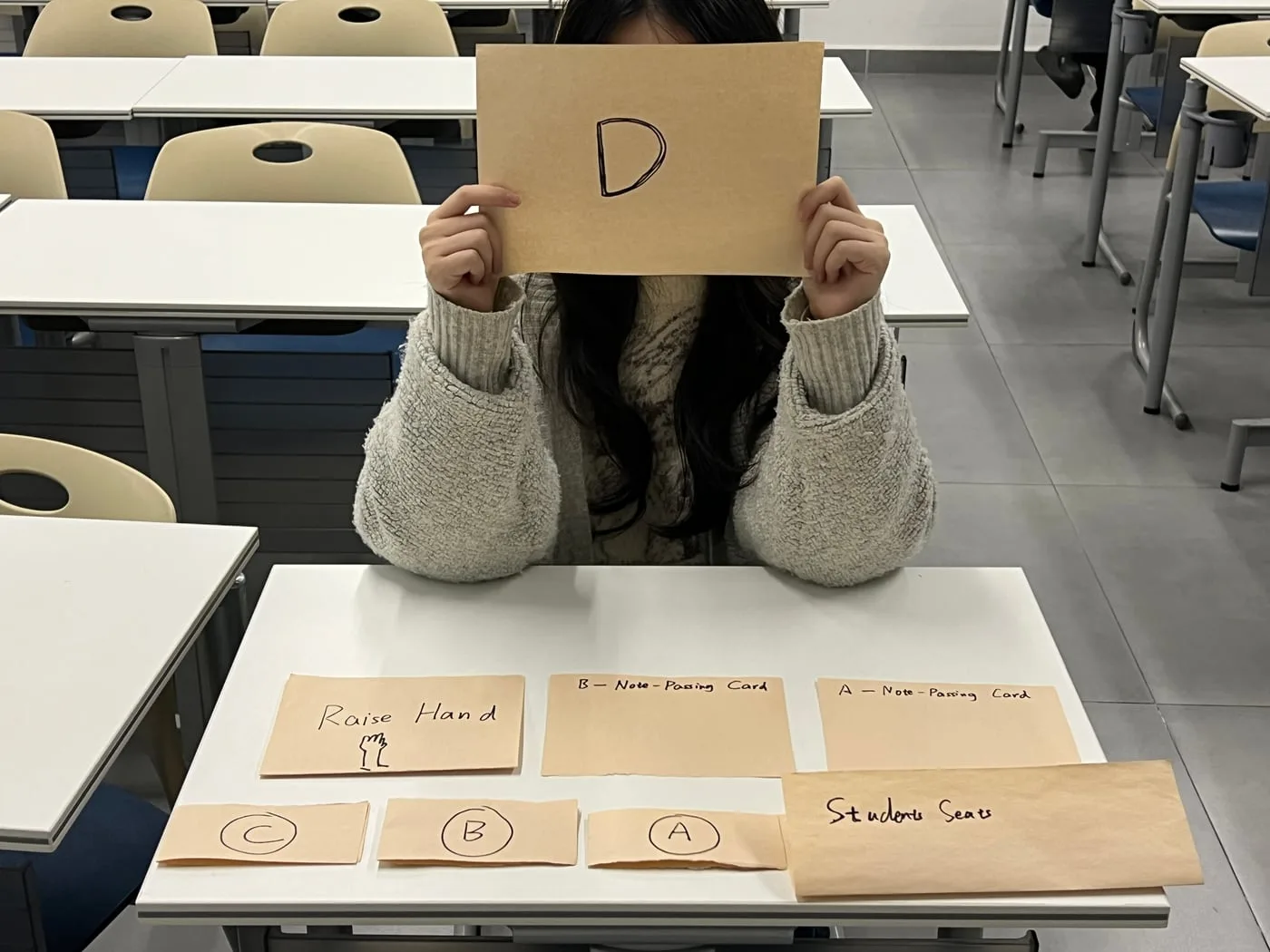 A student holding up a card marked D in front of their face, paper props on the desk