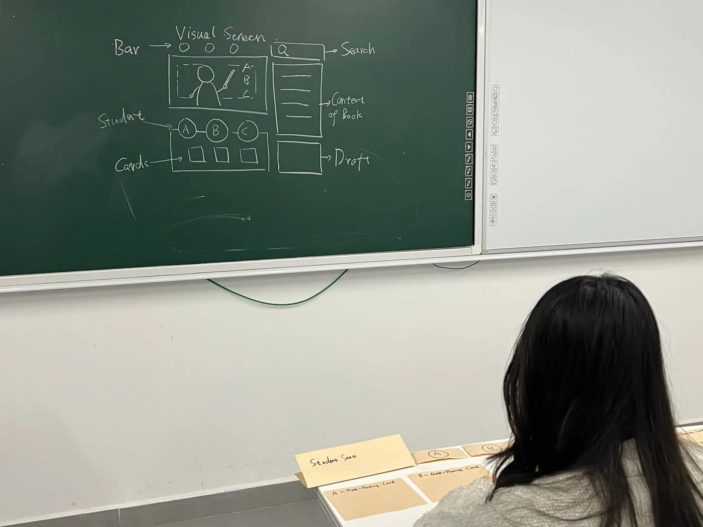 A student seated facing a chalkboard drawing of a virtual classroom screen, paper cards on the desk