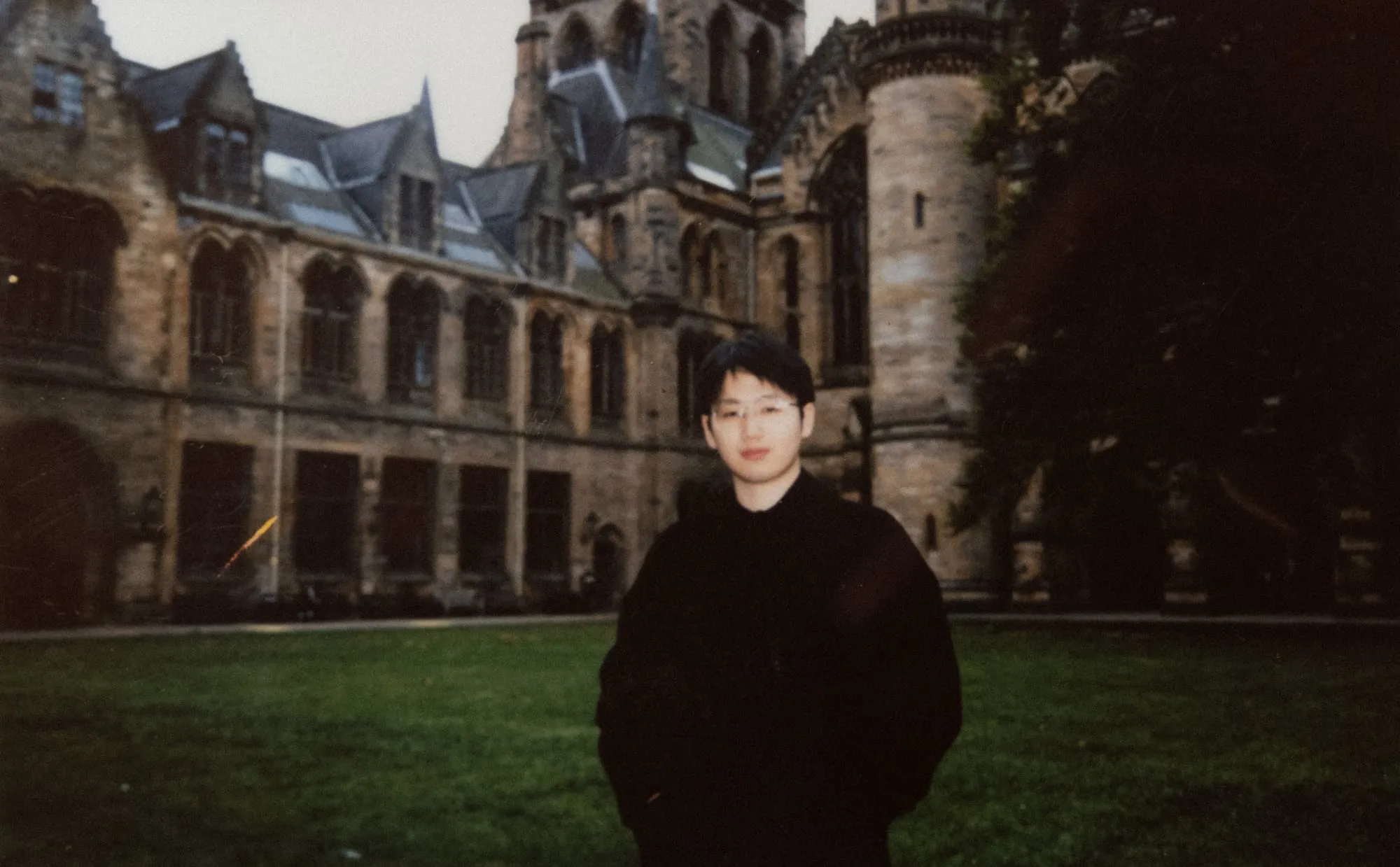 Wu Yiyang in a dark coat on a lawn in front of a Gothic sandstone quadrangle in Glasgow, on instant film