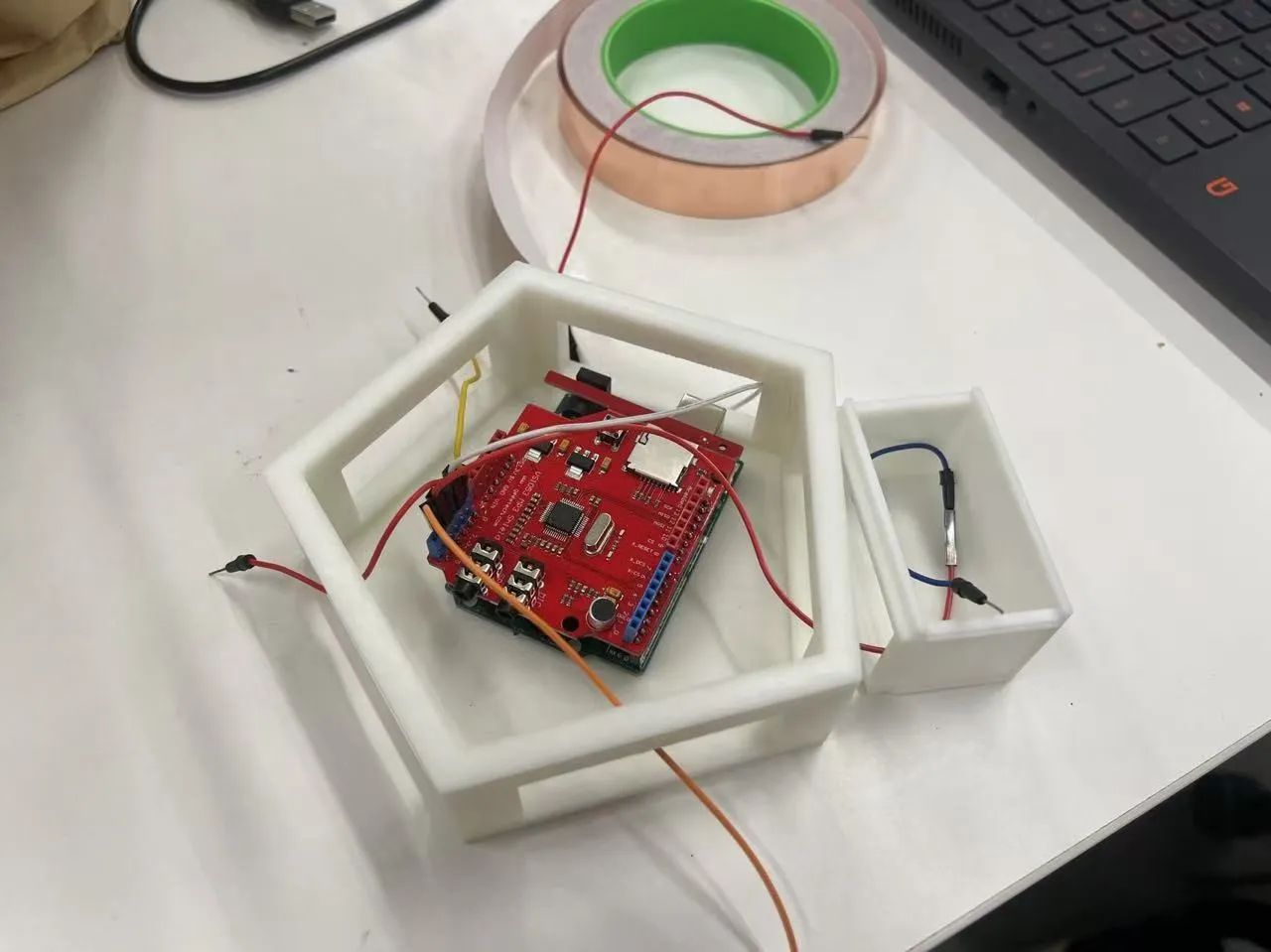 A 3D-printed white pentagon shell with a red shield board on an Arduino inside and leads running to one module