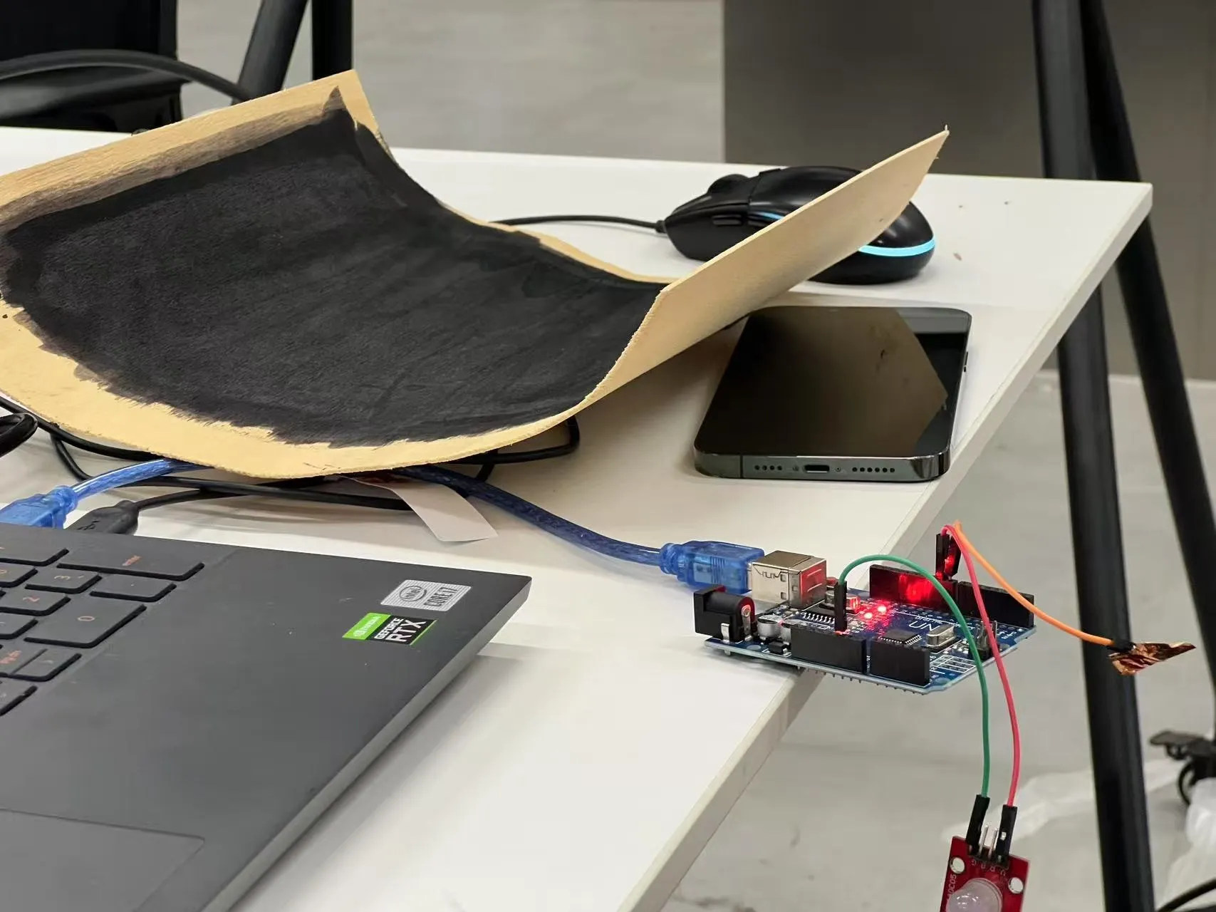 A large sheet of processed, darkened wood wired to an Arduino on a desk beside a laptop