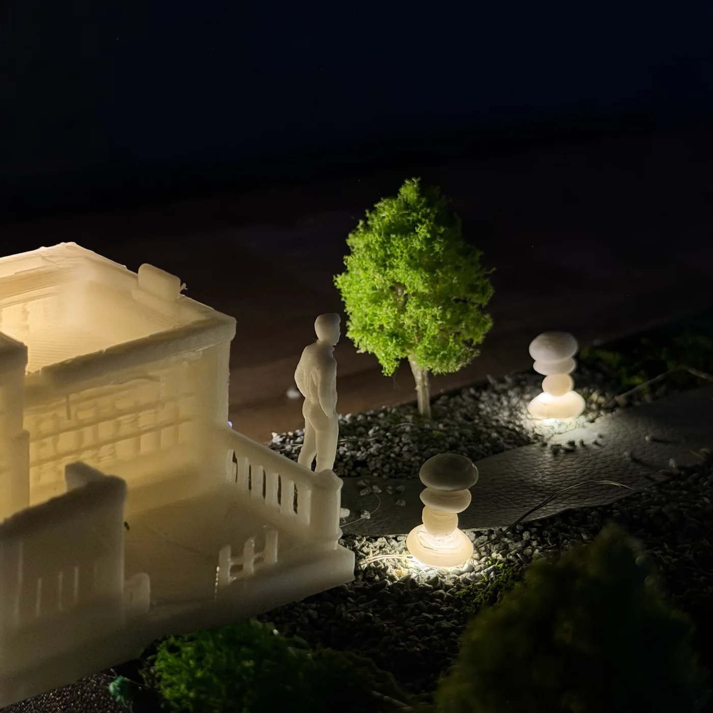 Close view of the site model: a figure beside a lit cairn on the model path, a white building behind
